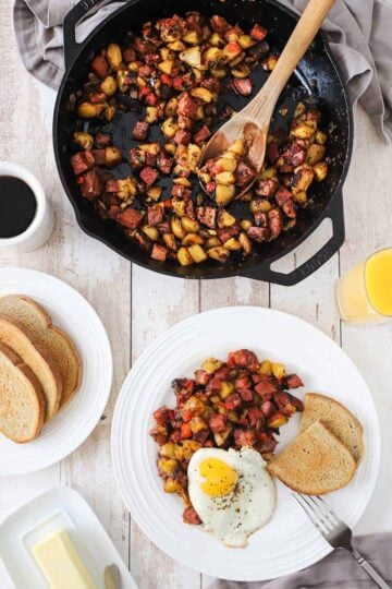 An overhead view of a white plate filled with a serving of corned beef hash with a fried egg on top and two slices of rye bread next to it as well as a skillet of the hash near the plate of food.