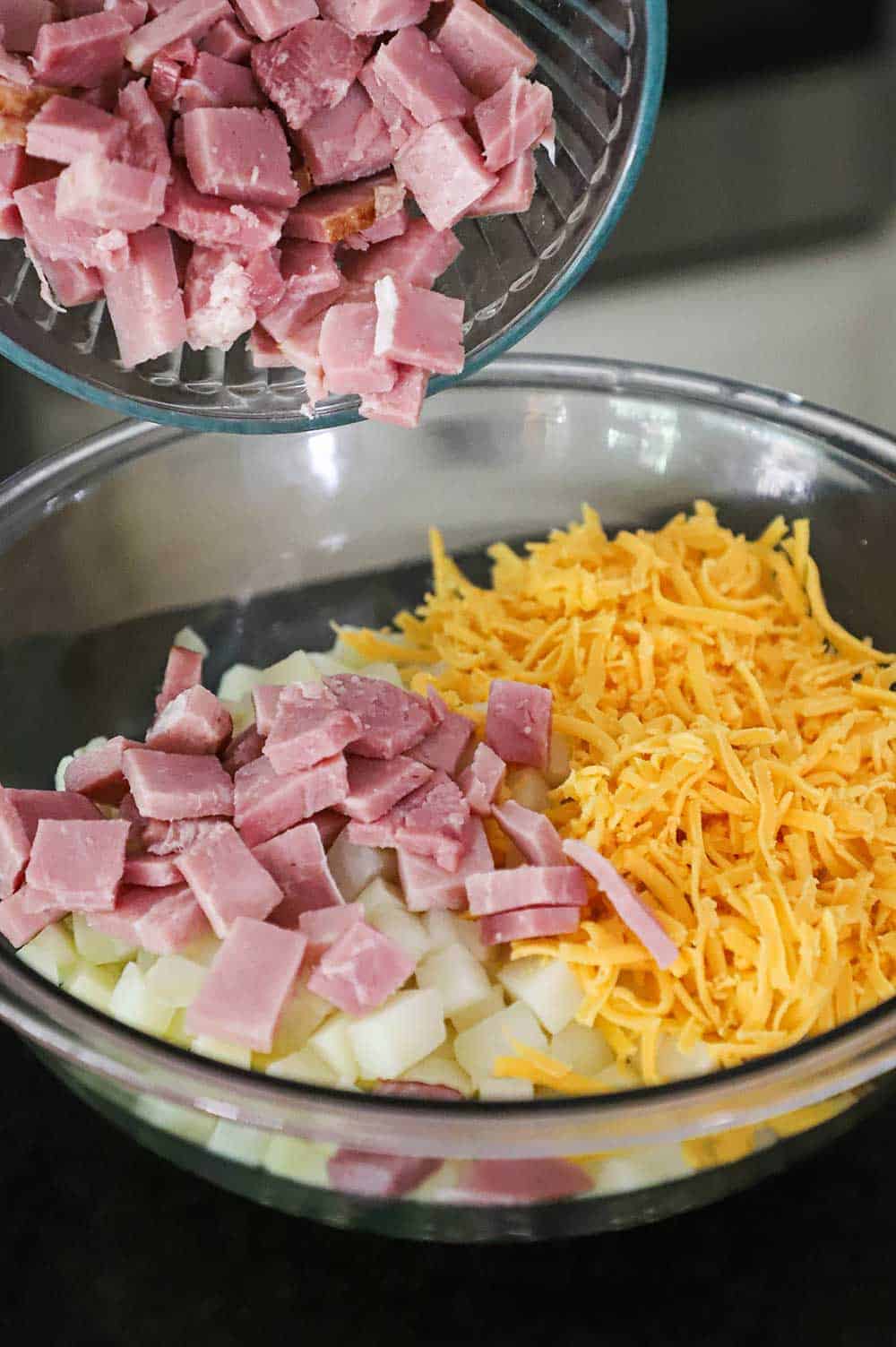 Chunks of ham being dumped from one glass bowl into a large glass bowl that is filled with a pile of cubed potatoes and topped with shredded cheddar cheese.