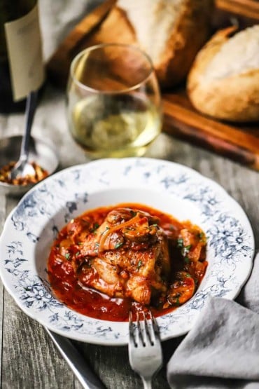 A straight-on view of a vintage individual bowl filled with a helping of chicken cacciatore and sitting next to a stemless glass of white wine.