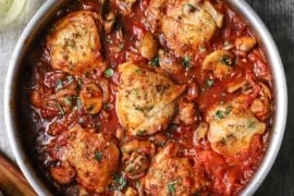 An overhead view of a large silver skillet filled with chicken cacciatore consisting of six seared chicken thighs in a thick tomato sauce.