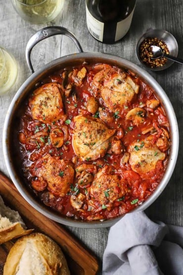 An overhead view of a large silver skillet filled with chicken cacciatore consisting of six seared chicken thighs in a thick tomato sauce.