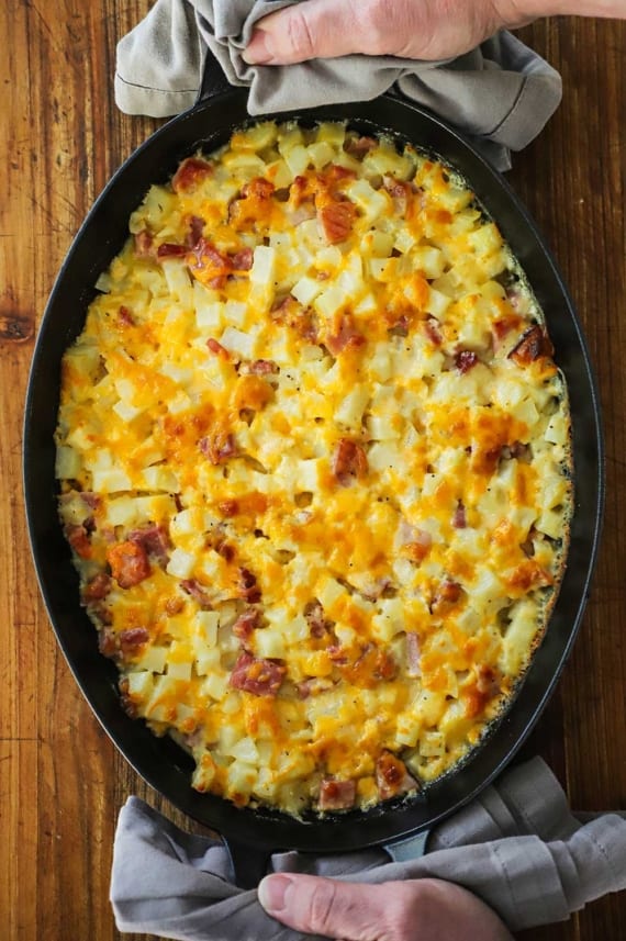 An overhead view of a person using two grey napkins to hold an oval baking dish that is filled with a freshly baked cheesy ham and potato casserole.