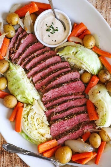 An overhead view of a large white oval platter that is filled with sliced corned beef flanked by sautéed cabbage, carrots, and baby yellow potatoes.