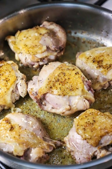 A close-up view of six chicken thighs with crispy skins being seared in a silver skillet in a thin layer of oil.