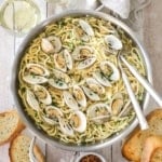An overhead view of a large stainless steel skillet filled with spaghetti all vongole with opened clams resting on top of the cooked pasta and two serving utensils inserted into the side of the dish.