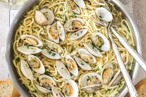 An overhead view of a large stainless steel skillet filled with spaghetti all vongole with opened clams resting on top of the cooked pasta and two serving utensils inserted into the side of the dish.