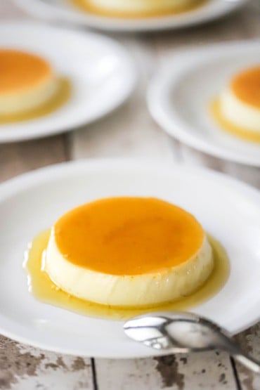 A straight-on view of homemade flan that has a light caramel sauce on top of it and surrounding the base and is on a white plate with other plated servings nearby.