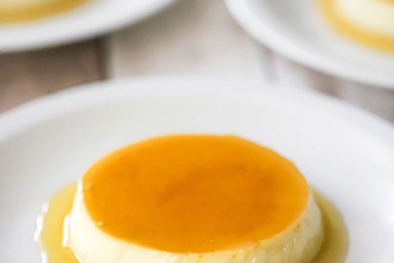 A straight-on view of homemade flan that has a light caramel sauce on top of it and surrounding the base and is on a white plate with other plated servings nearby.