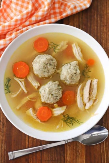 An overhead view of a shallow white soup bowl filled with homemade matzo ball soup with chicken and carrots and several sprigs of fresh dill.