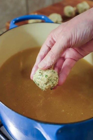A person holding an uncooked matzo ball over an oval Dutch oven that is filled with simmering chicken broth.