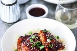 A straight-on view of two white dinner bowls filled with a serving of baked teriyaki chicken resting on a bed of steamed rice with a glass of white wine near by.