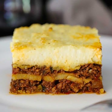 A straight-on view of a perfectly square piece of homemade moussaka sitting on a white dinner plate.