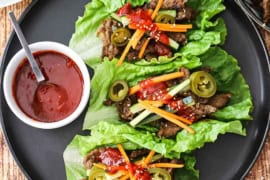 An overhead view of three beef bulgogi lettuce wraps that are sitting on a black plate and next to a small bowl filled with Korean barbecue sauce.