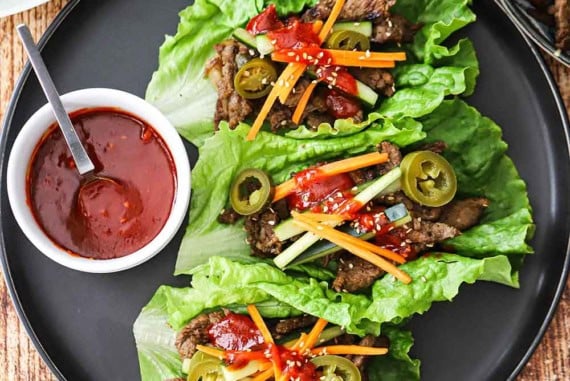 An overhead view of three beef bulgogi lettuce wraps that are sitting on a black plate and next to a small bowl filled with Korean barbecue sauce.