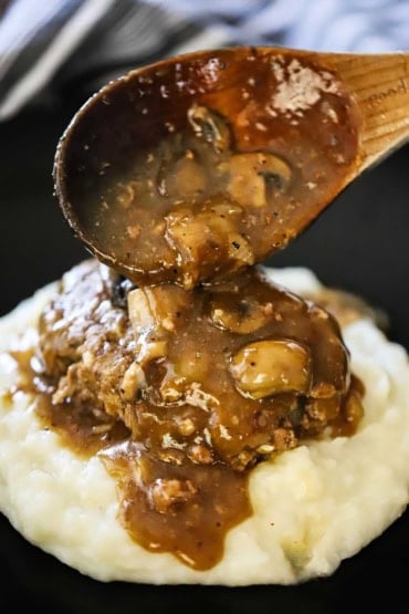 A large wooden spoon being used to pour a mushroom brown gravy over a salisbury steak that is resting on pile of mashed potatoes.