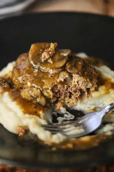 A close-up view of a half eaten salisbury steak with mushroom brown gravy over mashed potatoes on a black dinner plate.