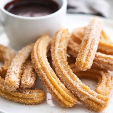 A close-up view of a stack of homemade churros sitting on a white plate next to a small cup filled with chocolate sauce.