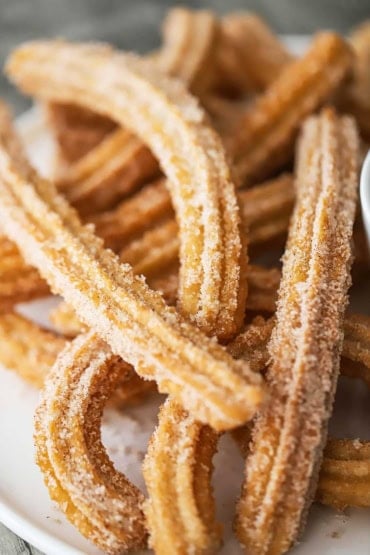 A close-up view of a pile of homemade churros stacked on a white circular platter.