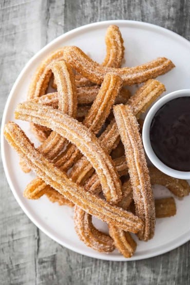 An overhead view of a circular white plate that is filled on one side with a pile of homemade churros and a small bowl of chocolate sauce on the other side.
