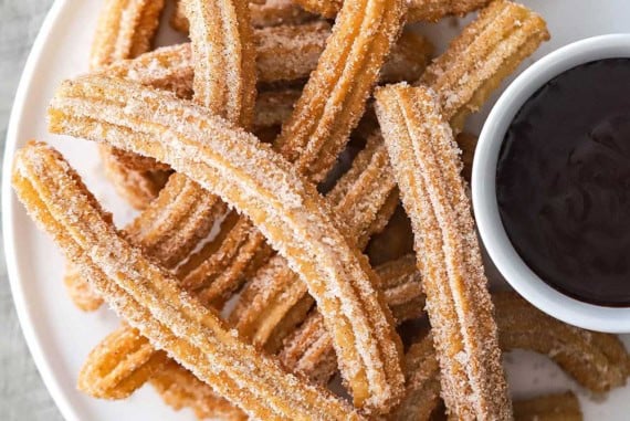 An overhead view of a circular white plate that is filled on one side with a pile of homemade churros and a small bowl of chocolate sauce on the other side.