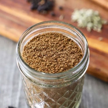 A close-up view of a small glass jar filled with freshly ground garam masala next to a cutting board topped with various spices.