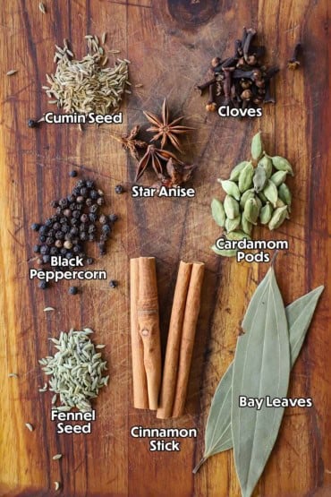 A overhead view of a variety of spices and aromatics sitting in small piles on a wooden cutting board. The name of the spice appears over each one.