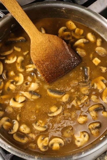 An overhead view of a large silver skillet filled with a brown gravy with sautéed sliced mushrooms with a wooden spatula in the middle of it.