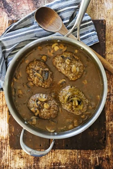 An overhead view of a large silver saucepan filled with four salisbury steaks that are resting in a mushroom brown gravy with a wooden spoon resting nearby.