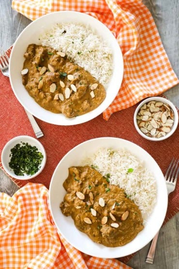 An overhead view of two white bowls filled with a serving of chicken korma next to a pile of basmati rice with small bowls of sliced almonds and chopped cilantro nearby.