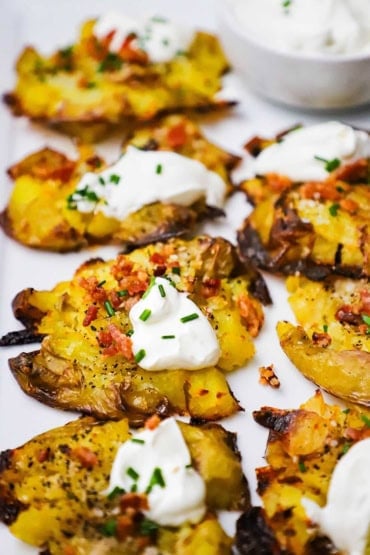 A close-up view of cooked smashed potatoes that are brown and crispy on the edges that have been topped with crumbled bacon, a dollop of sour cream, and snipped chives.