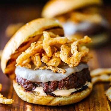 A straight-on view of a smoked steakhouse burger topped with melty provolone cheese and crispy onion strips and a layer of steak sauce aioli on the bottom bun.