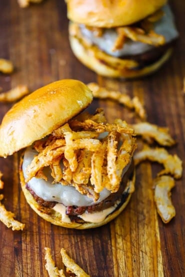 An overhead view of a smoked steakhouse burger sitting on a wooden cutting board with another similar burger in the background.