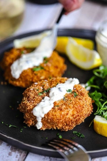 A straight-on view of two New England-style crab cakes that are on a black dinner plate both with a strip of tartar sauce on them and sitting next to lemon wedges.