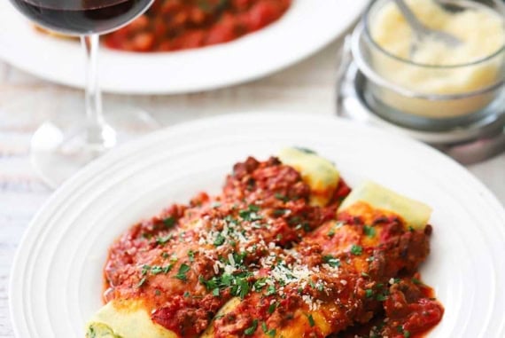 A straight-on view of a white dinner plate that is filled with two side-by-side spinach cannelloni and meat sauce near a wine glass partially filled with red wine.