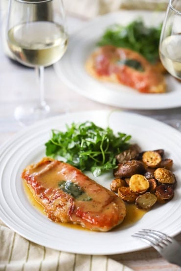 A straight-on view of a white dinner plate that is holding a serving of chicken saltimbocca with a white wine butter sauce next to roasted baby potatoes and an arugula salad.