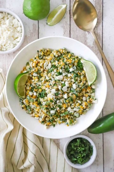 An overhead view of a white bowl that is filled with a large serving of Mexican street corn salad (equites) with a small bowl of crumbled cheese and a fresh jalapeño sitting nearby.