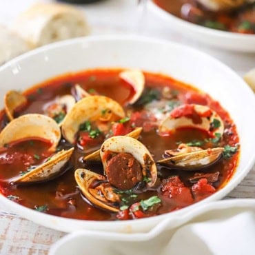 A close-up view of a white bowl filled with a serving of Portuguese-style clam chowder with pieces of cooked chorizo, tomato chunks, and opened littleneck clams visible.