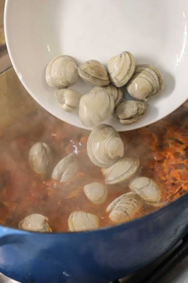 A person dumping uncooked littleneck clams from a shallow white bowl into a large Dutch oven filled with a simmering tomato-based broth.