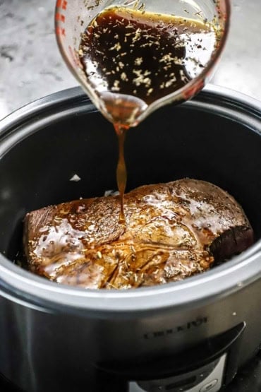 A person pouring a beef broth mixture with herbs over a seared chuck roast that is resting inside a large slow cooker.