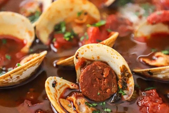 A close-up view of a white bowl filled with a serving of Portuguese-style clam chowder with pieces of cooked chorizo, tomato chunks, and opened littleneck clams visible.