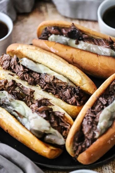 A straight-on view of four large slow cooker French dip sandwiches resting on a black plate with two small bowls filled with au jus sauce nearby.