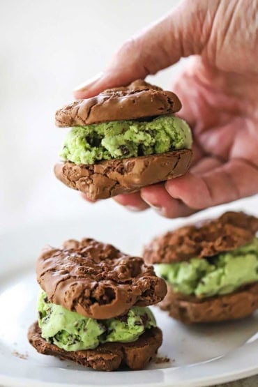 A person holding a mint chocolate chip ice cream sandwich with the ice cream sandwiches between two double chocolate chip cookies.