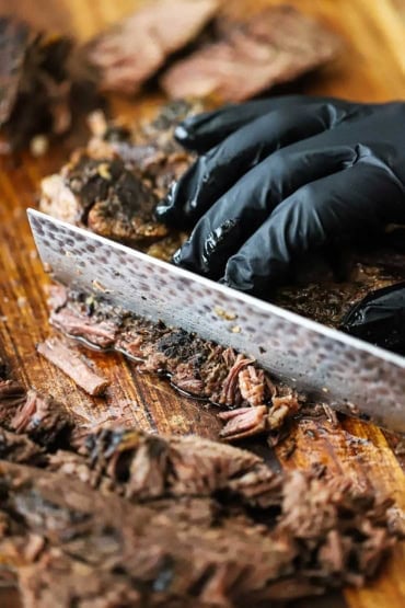 A person wearing a black glove holding a slow-cooked chuck roast and slicing it into juicy strips with a large Japanese chef's knife.