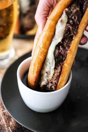 A person plunging a whole slow cooker French dip sandwich in a small white bowl filled with au jus sauce sitting on a black circular plate.