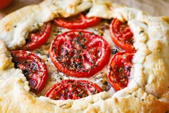 A straight-on view of a baked tomato galette with gruyere cheese and caramelized onions sitting next to a sprig of thyme and two tomatoes on a vine.
