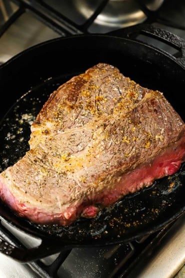 A large chuck roast being seared in a large black cast-iron skillet and is lightly browned on the top surface of the meat.