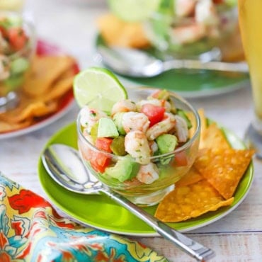 A straight-on view of a small glass bowl that is filled with a serving of Mexican shrimp ceviche and is on a green cocktail plate with corn tortilla chips and a silver spoon on it.