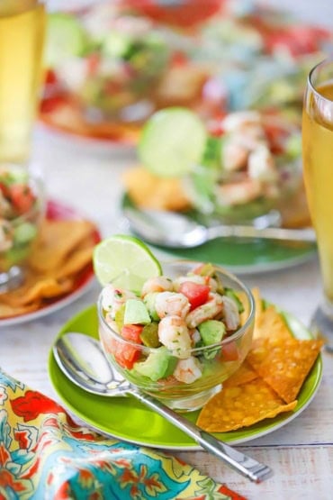 A straight-on view of a small glass bowl that is filled with a serving of Mexican shrimp ceviche and is on a green cocktail plate with corn tortilla chips and a silver spoon on it.