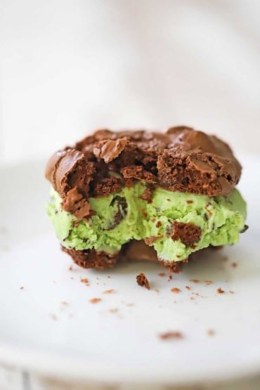 A close-up view of a mint chocolate chip ice cream cookie sandwich with a bite take out of it and sitting on a white dessert plate.