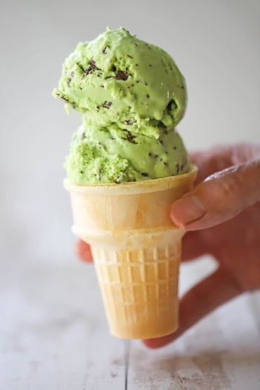 A person holding an old-fashioned ice cream cone with two large scoops of mint chocolate chip ice cream piled on top.